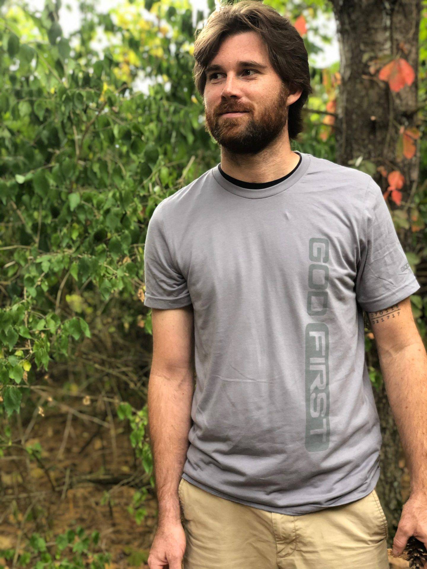 Man in forest with tattoos and has a beard and long hair with a grey shirt that says God first.