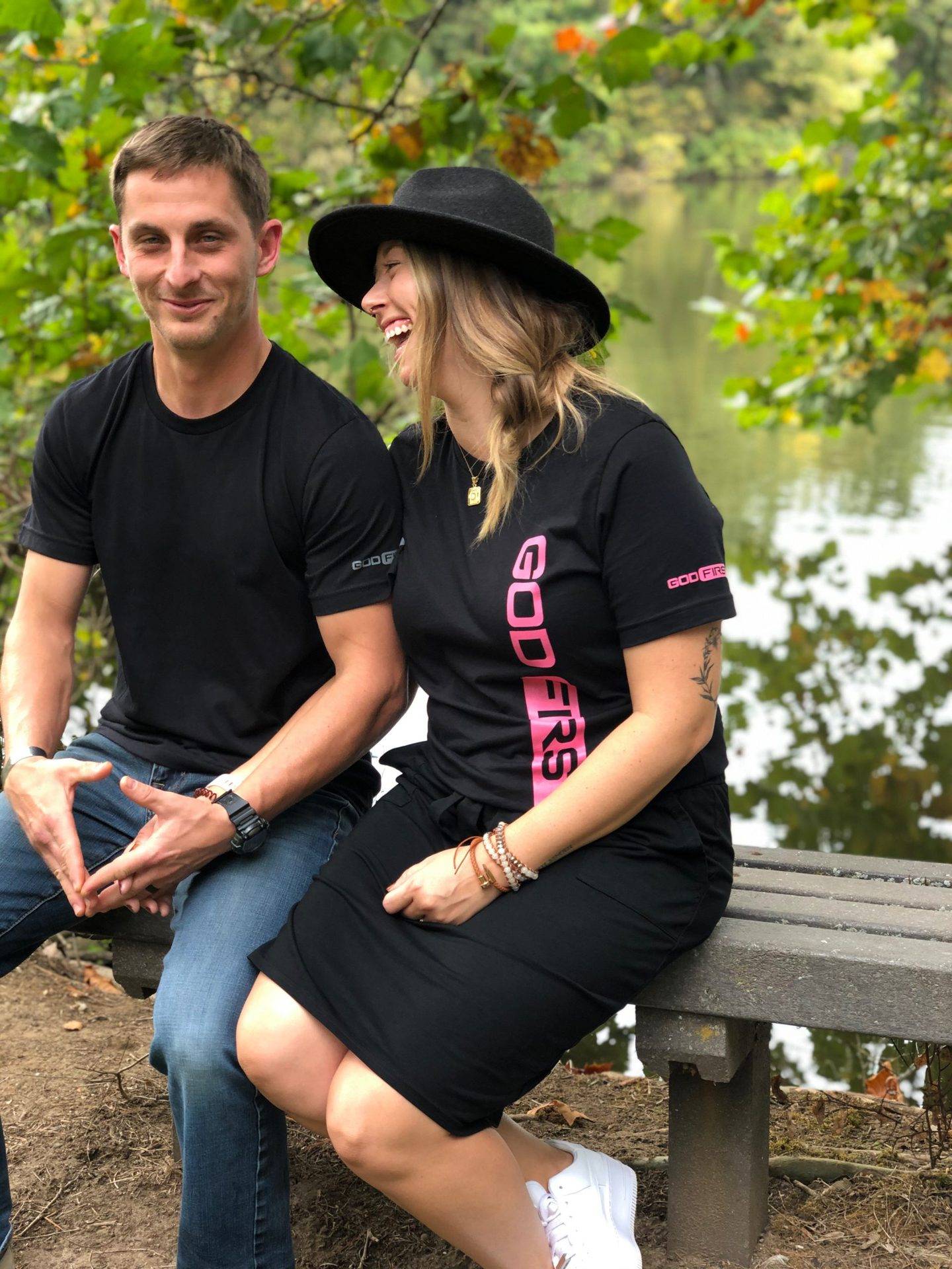 Man and women laughing the woman has long hair and a hat and they are both wearing a black shirt in a forest sitting.