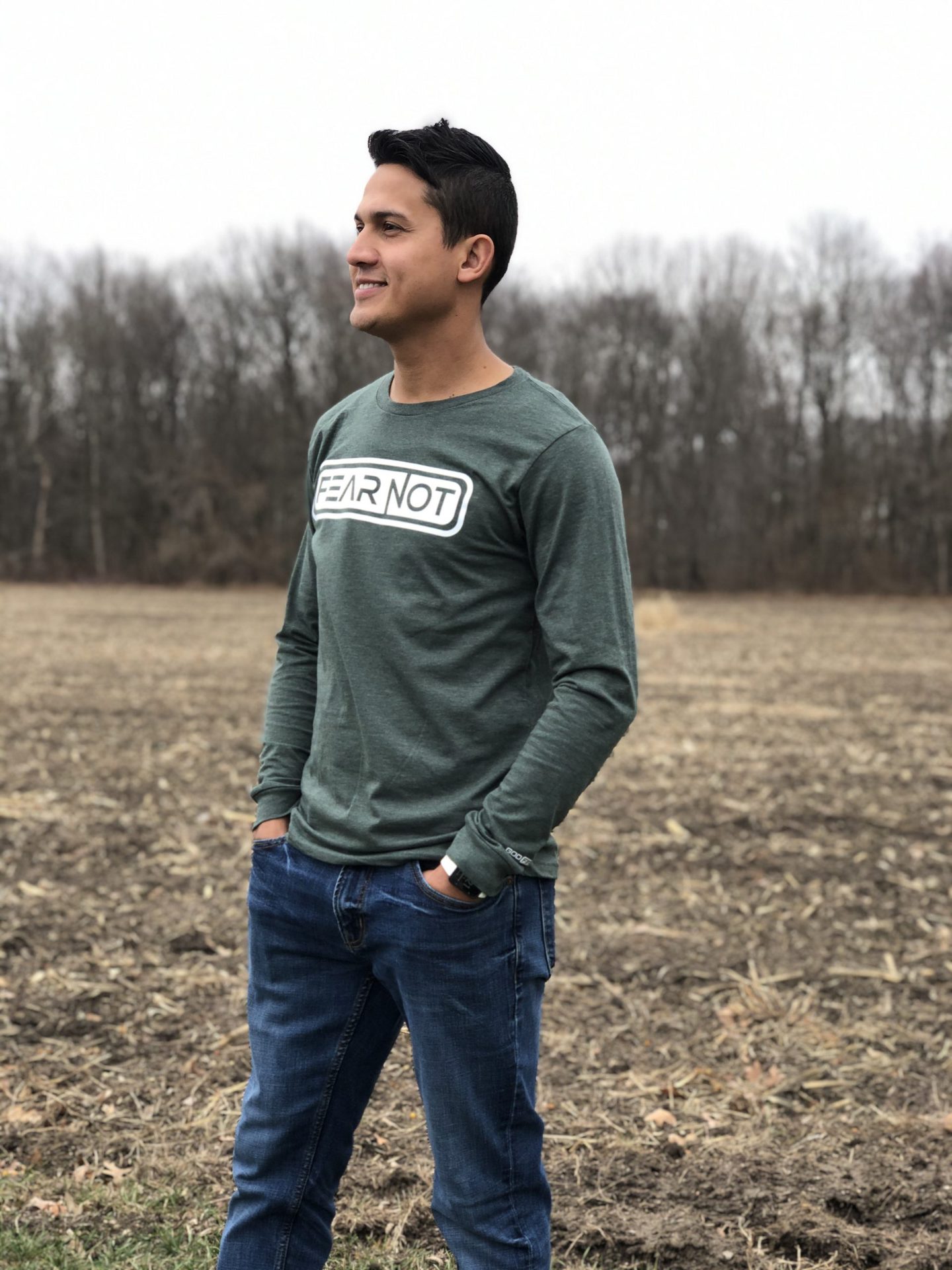 Man in Forest color shirt with hands in pockets in a field the shirt says Fear not.