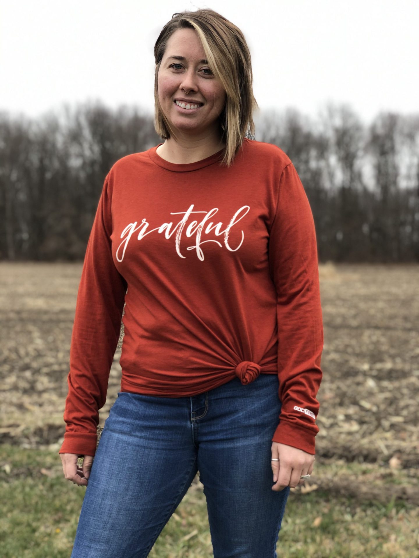 Women in a field with her shirt tied and says grateful long hair and brick colored shirt.